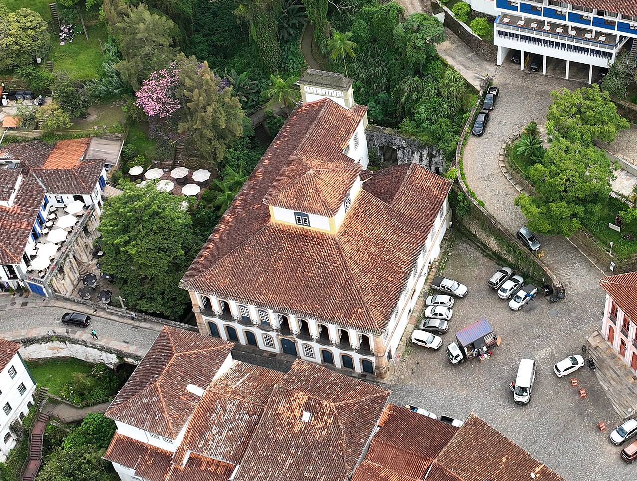 Vista aérea da Casa dos Contos, sobrado claro com detalhes ocres, portas azuis e telhados de barro, cercado por ruas e casario.