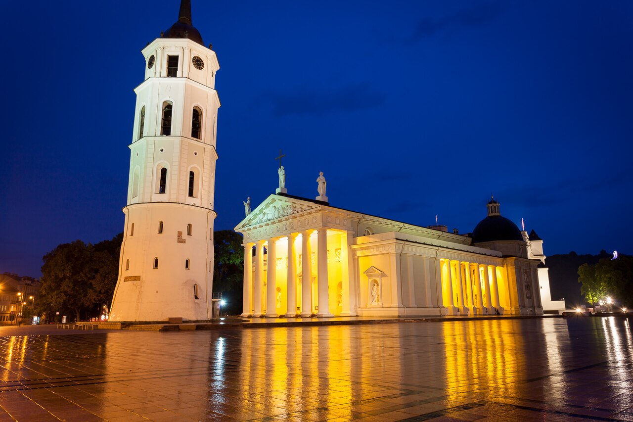 Catedral branca iluminada ao anoitecer, com colunas na fachada, campanário separado à esquerda e reflexos na praça molhada.