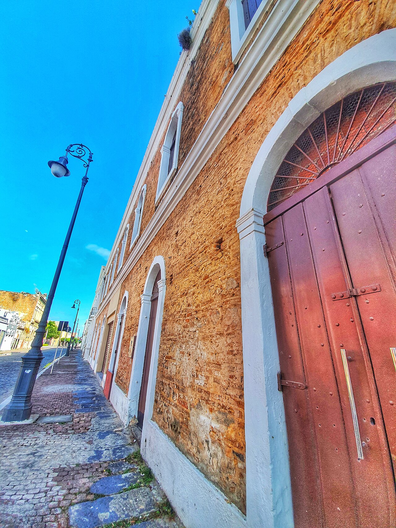Rua de pedras em Jaraguá com postes ornamentais e edifício de alvenaria aparente, portas altas em arco e molduras brancas.