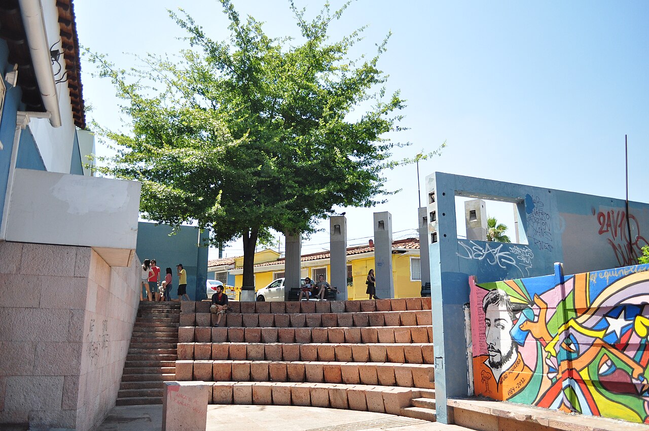Área externa de La Chascona com degraus de pedra, uma árvore, pessoas, construção azul e branca e mural colorido com retrato masculino.