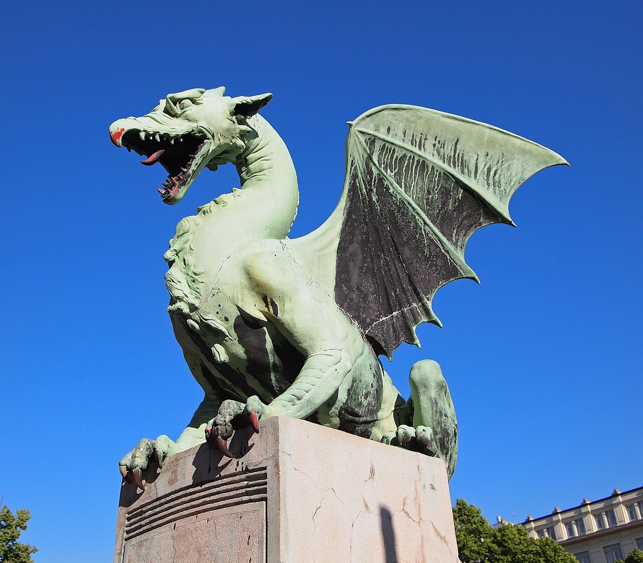Escultura esverdeada de dragão com boca aberta, garras e asas sobre um pedestal de pedra, com árvores e céu azul ao fundo.