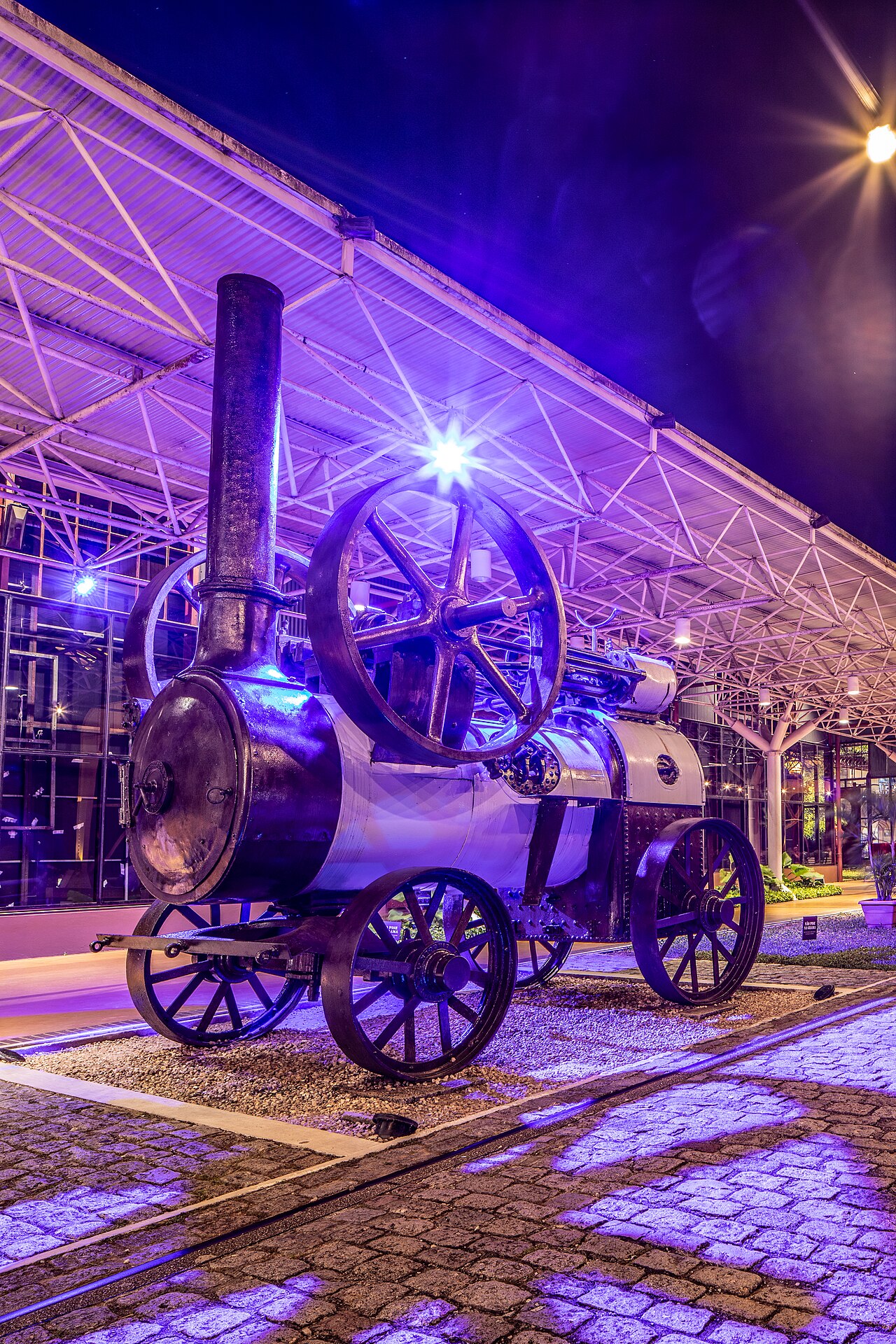 Máquina a vapor histórica com chaminé alta e grande volante sob estrutura metálica, iluminada em roxo e azul na Estação das Docas.