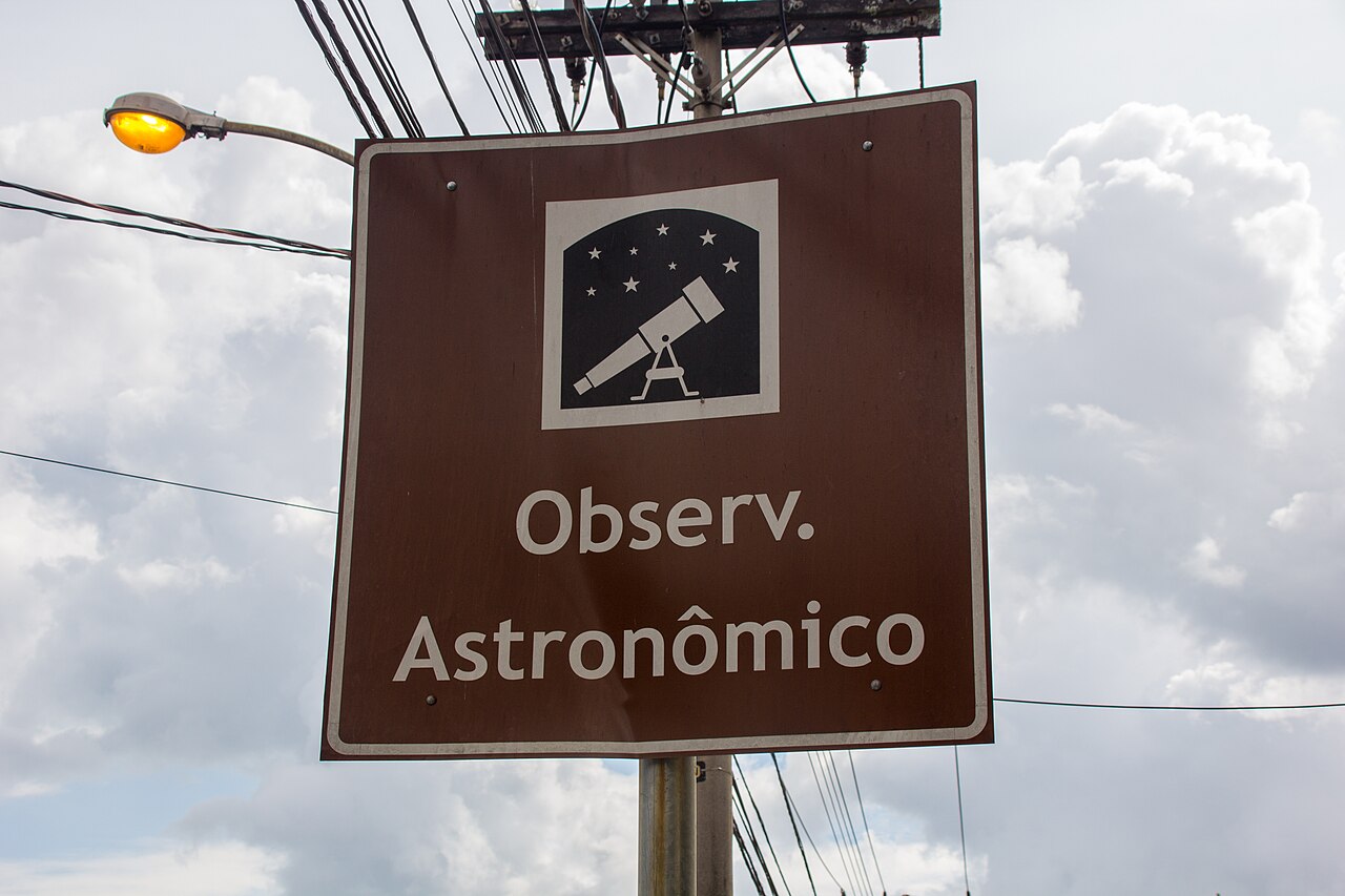Placa marrom com a inscrição Observ. Astronômico e pictograma de telescópio e estrelas, cercada por postes e fios sob céu com nuvens.