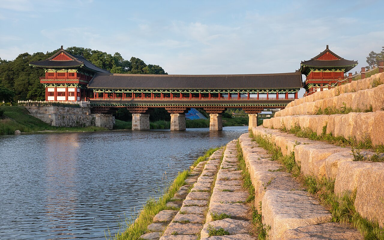 Ponte coberta Woljeonggyo com estrutura vermelha e detalhes verdes sobre pilares de pedra, vista da margem com degraus iluminados pelo sol.