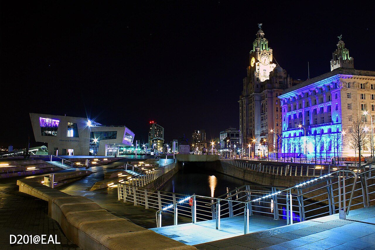 Pier Head à noite, com canal em primeiro plano, escadarias e corrimãos iluminados, terminal Mersey Ferries à esquerda e edifício monumental iluminado à direita.