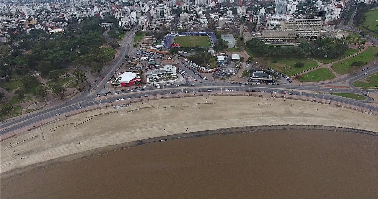 Vista aérea da região do Parque Rodó, com água amarronzada, faixa de areia, avenida costeira, árvores, brinquedos e prédios ao fundo.
