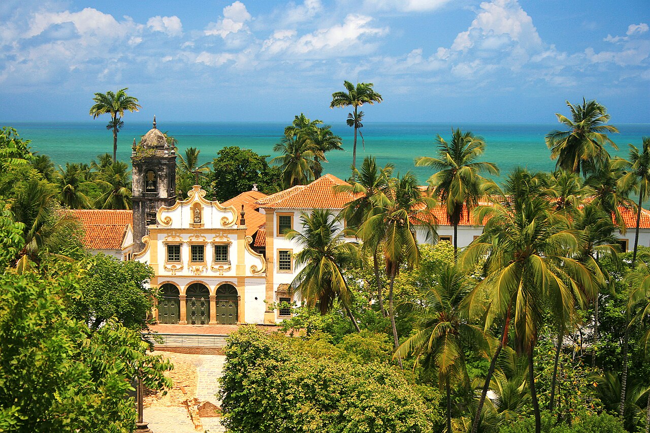 Convento de São Francisco com fachada branca e ocre, entradas em arco, torre escura e telhados cerâmicos, cercado por vegetação, com o mar ao fundo.