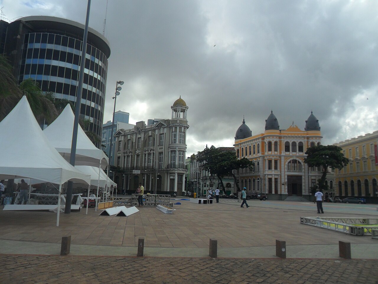 Praça do Marco Zero com pedestres, tendas brancas e estruturas metálicas diante de edifícios históricos, sob céu encoberto.