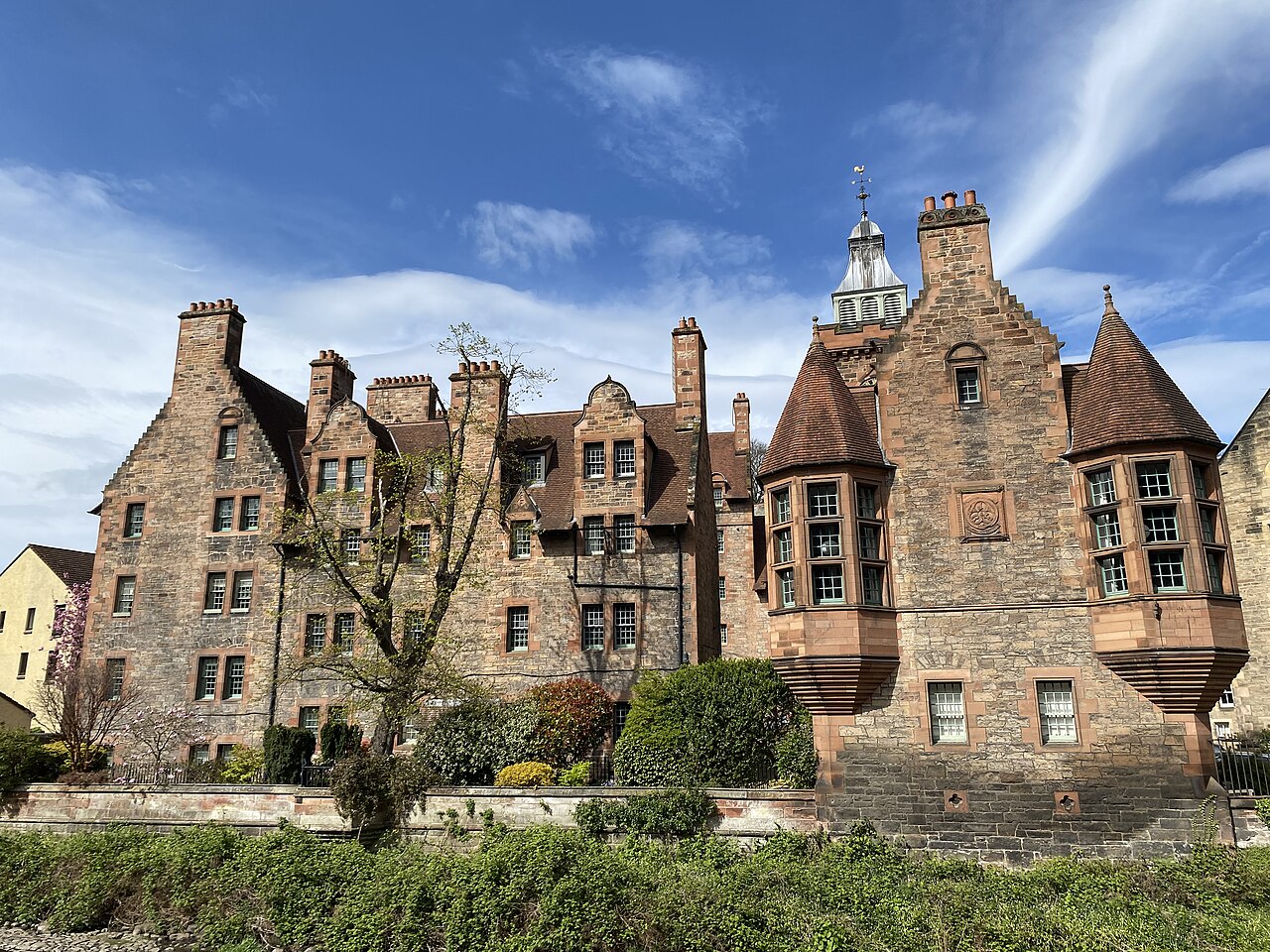Edifícios de pedra em Dean Village com empenas escalonadas, chaminés altas, torres de telhados cônicos e vegetação em primeiro plano.