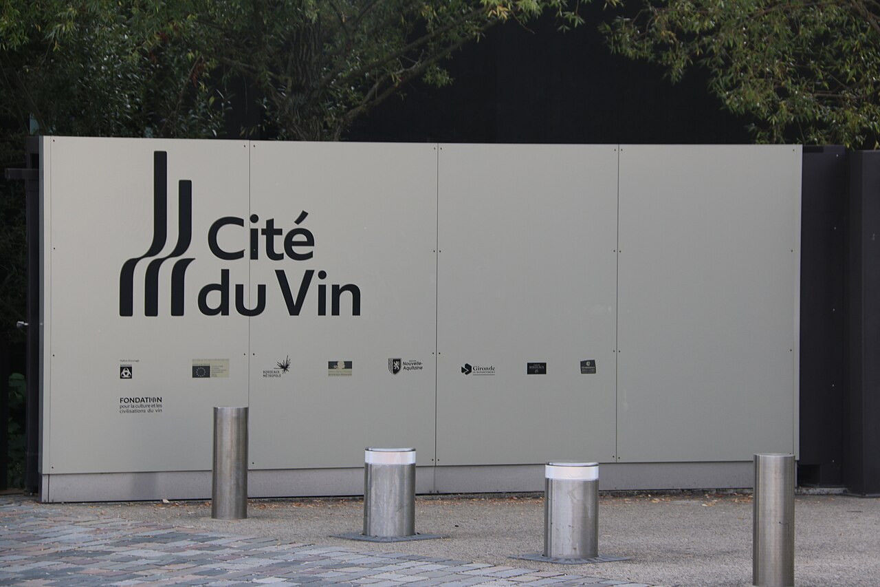 Painel claro com nome e logotipo da Cité du Vin, quatro balizadores metálicos à frente e vegetação ao fundo.