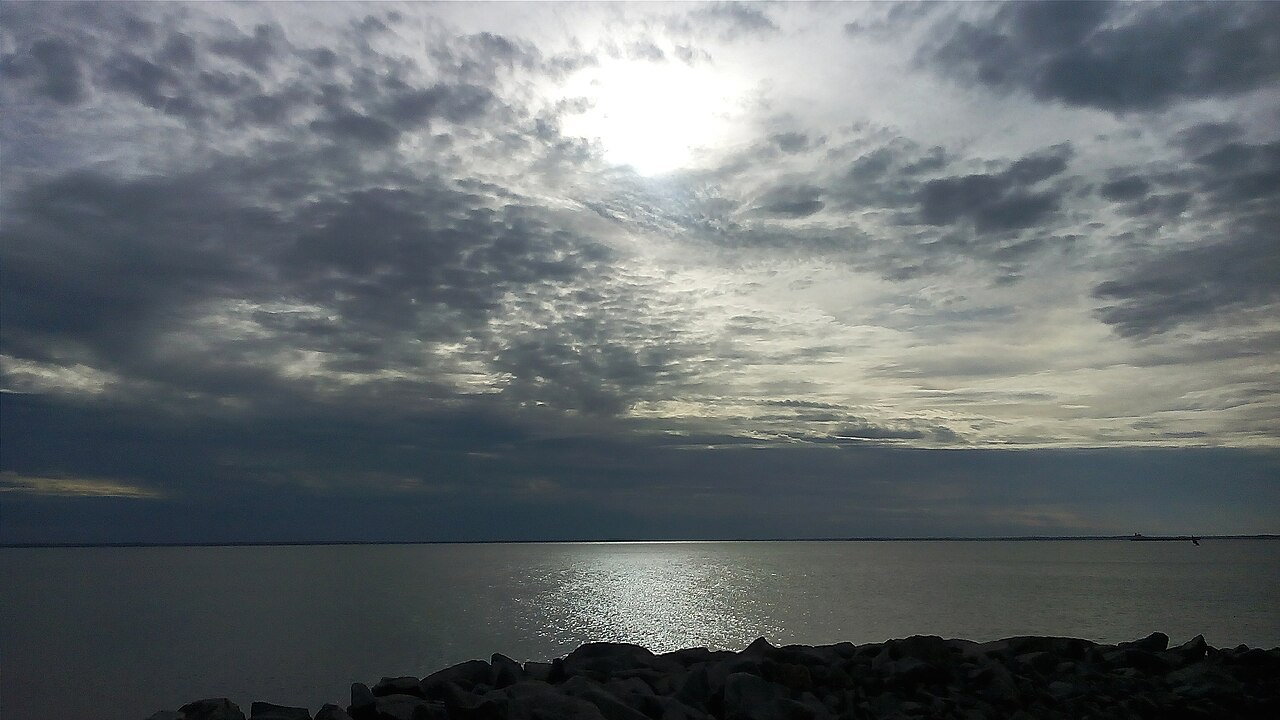 Vista de água calma e horizonte baixo a partir do Espigão Costeiro, com pedras escuras em primeiro plano e céu cheio de nuvens.