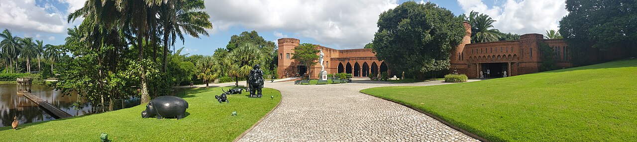 Panorama do Instituto Ricardo Brennand com edifícios de tijolos avermelhados, torres, caminho de pedras, gramados e lago à esquerda.