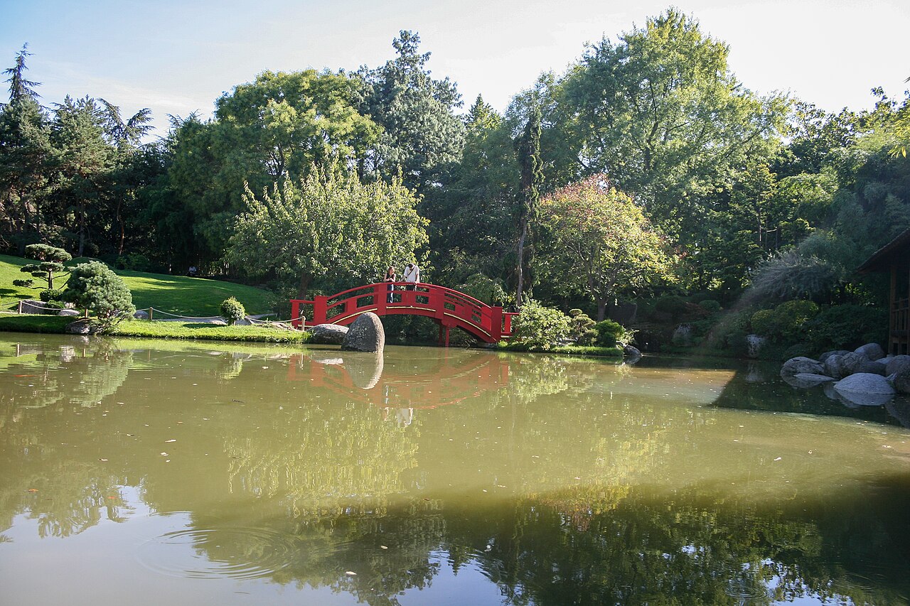 Ponte vermelha arqueada com duas pessoas sobre um lago, cercada por árvores, arbustos e pedras no Jardin japonais.