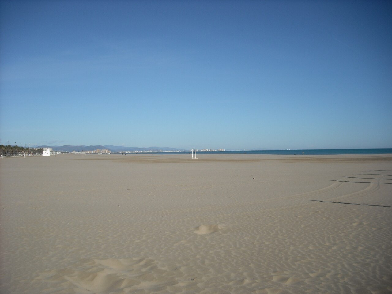 Faixa ampla de areia clara em Malvarrosa, quase vazia, com postes espaçados, mar azul e palmeiras à esquerda.