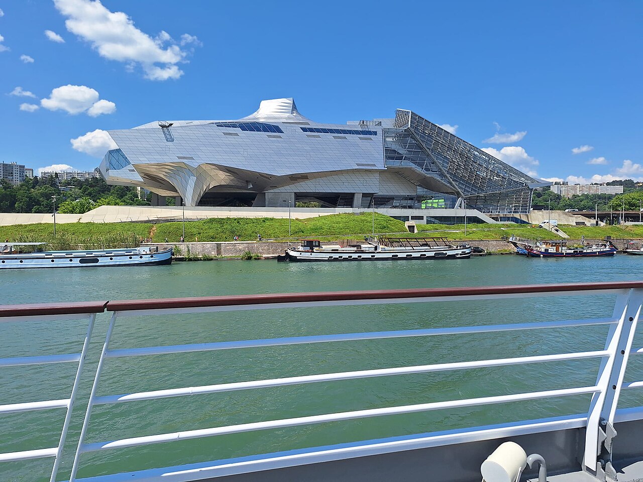 Musée des Confluences visto do Rhône, com volumes metálicos angulares, estrutura envidraçada e barcos junto à margem.