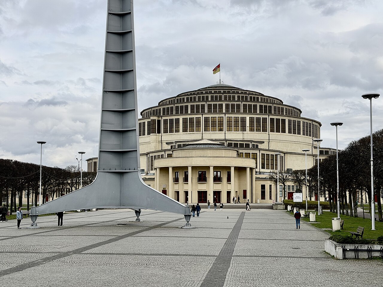 Hala Stulecia com cobertura circular escalonada e entrada com colunas, parcialmente encoberta por uma alta estrutura metálica de três apoios, diante de praça com pedestres.