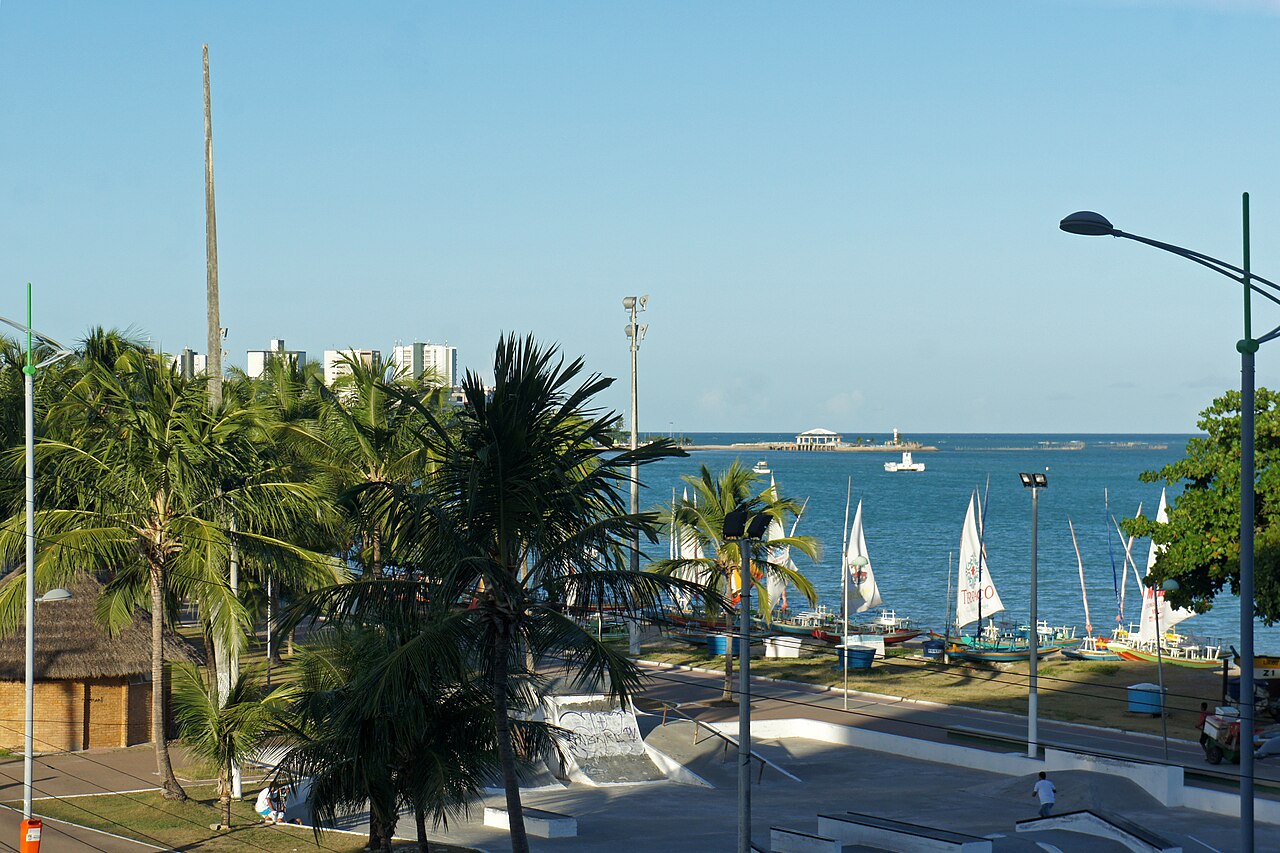 Orla de Pajuçara com pista de skate, coqueiros, avenida e jangadas de velas junto à margem.