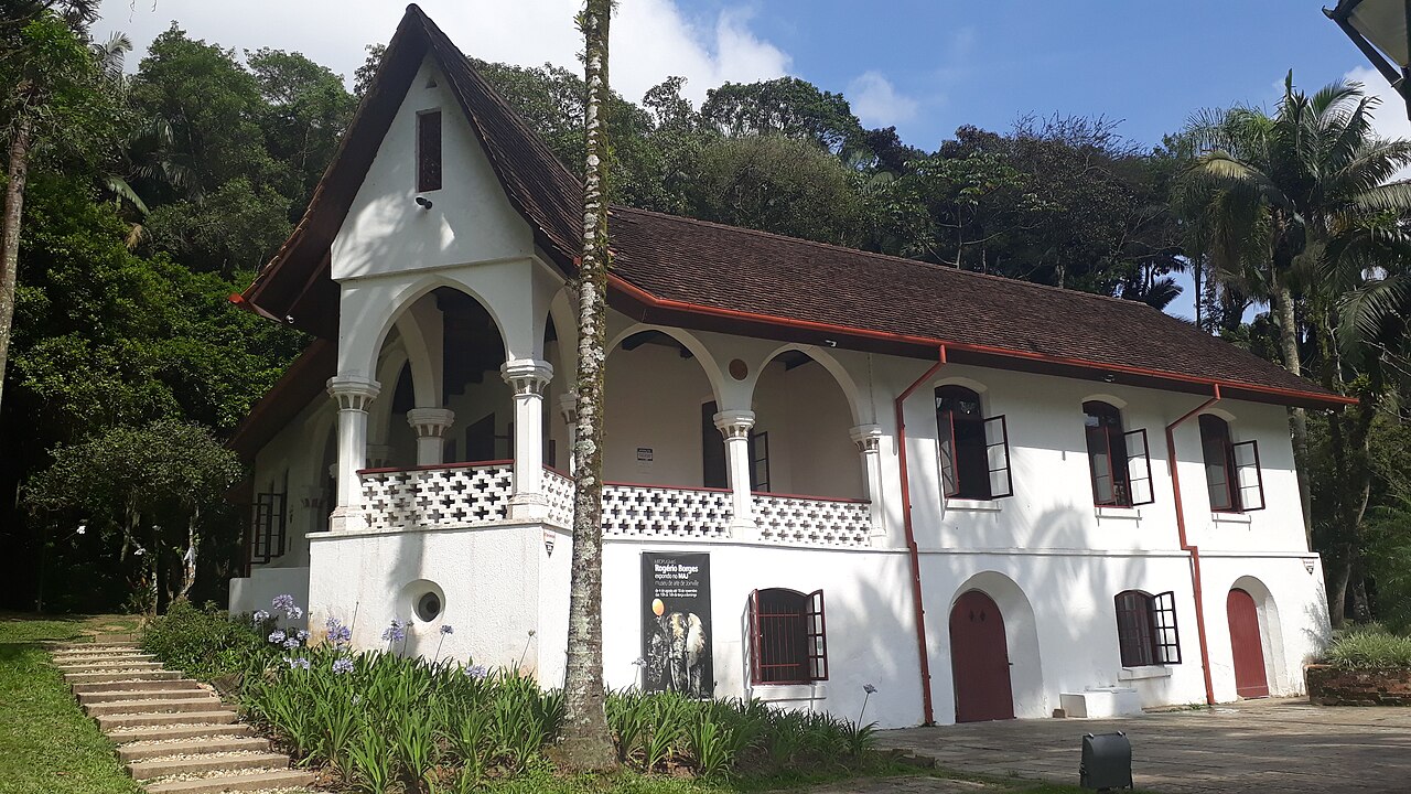Casarão branco de dois pavimentos, telhado escuro inclinado, varanda com arcos e janelas avermelhadas, cercado por jardim e vegetação.