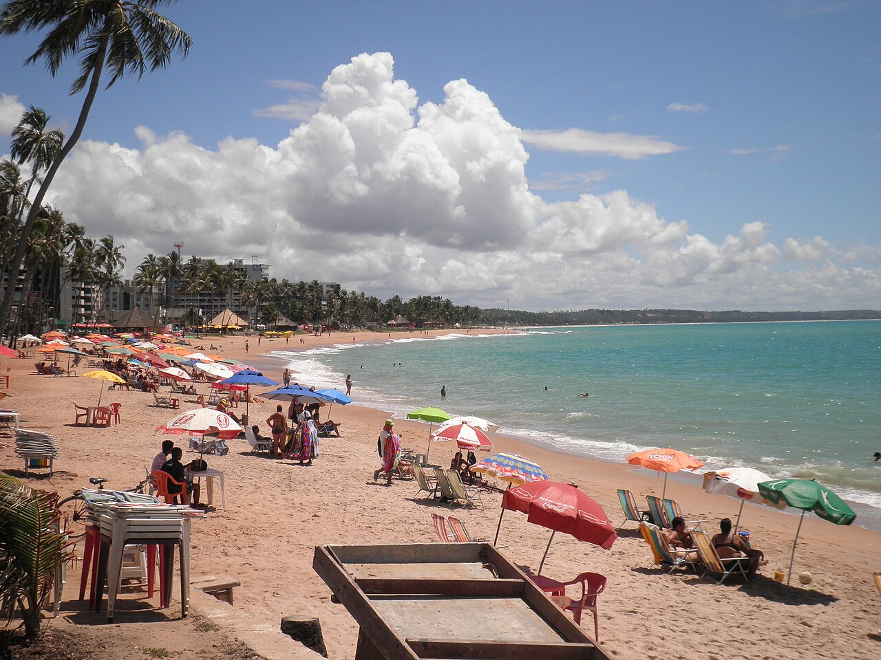 Praia de Ponta Verde com banhistas, guarda-sóis coloridos, coqueiros e edifícios ao longo da areia.