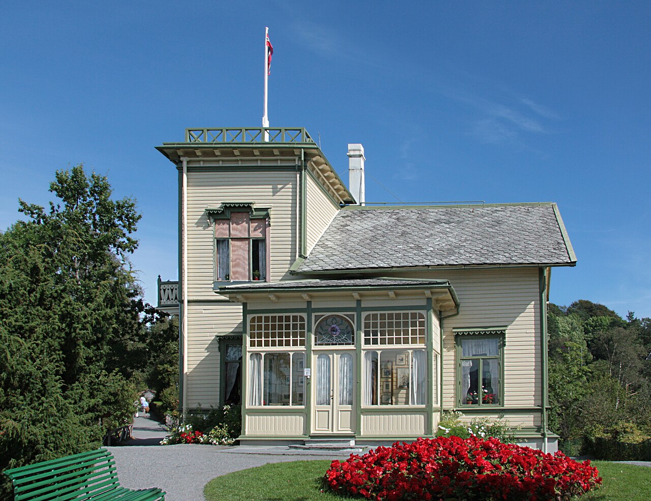 Casa de madeira clara com detalhes verdes, torre e varanda envidraçada, cercada por árvores e jardim em Troldhaugen.