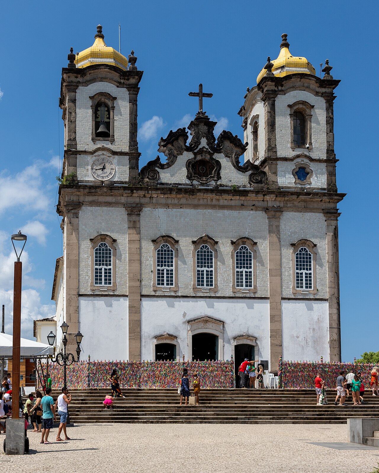 Fachada branca da Igreja do Bonfim com duas torres de cúpulas amarelas, fitinhas coloridas nas grades e pessoas na escadaria.