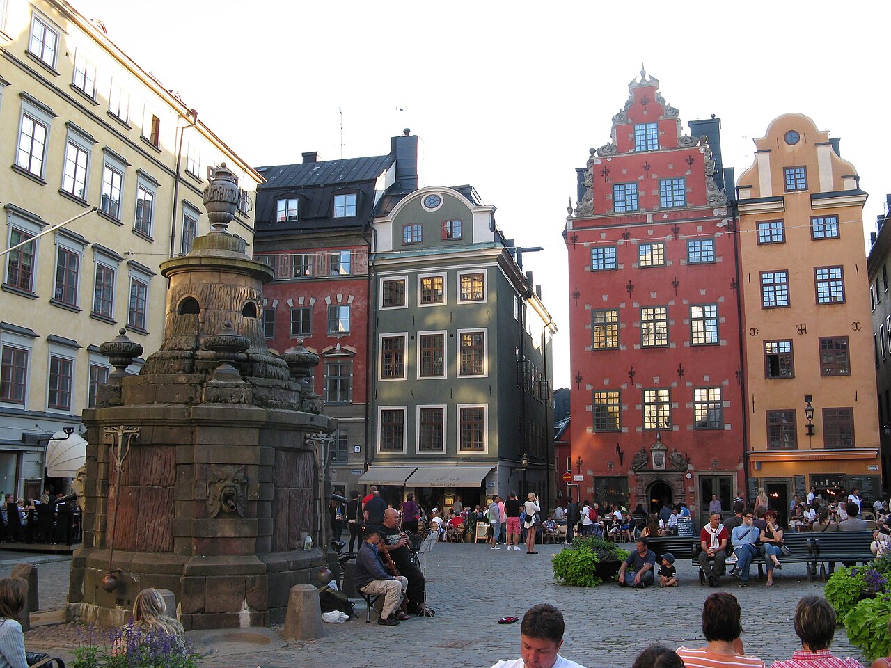 Praça Stortorget com calçamento de pedra, fonte à esquerda, fachadas históricas coloridas e pessoas.