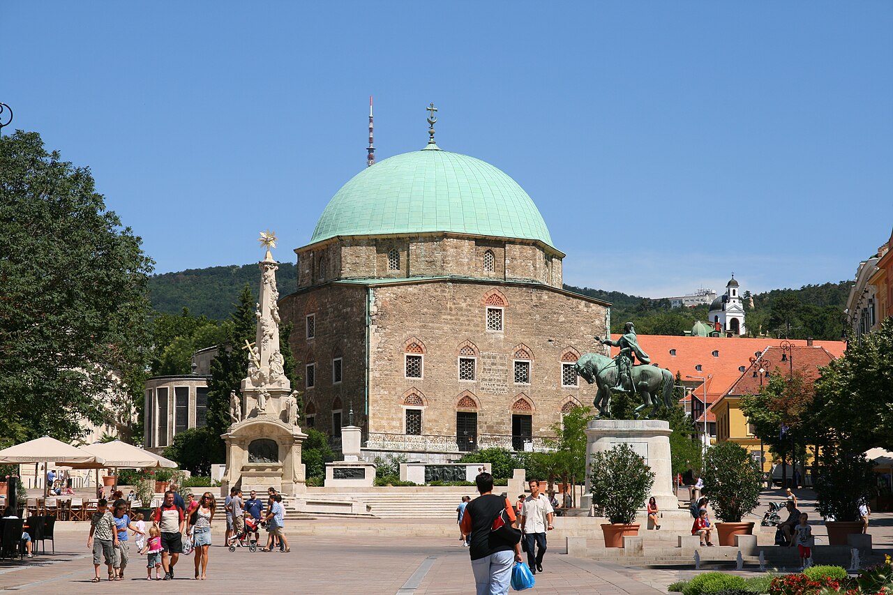 Praça Széchenyi com pedestres, edifício de cúpula verde, coluna ornamentada e estátua equestre sobre pedestal.