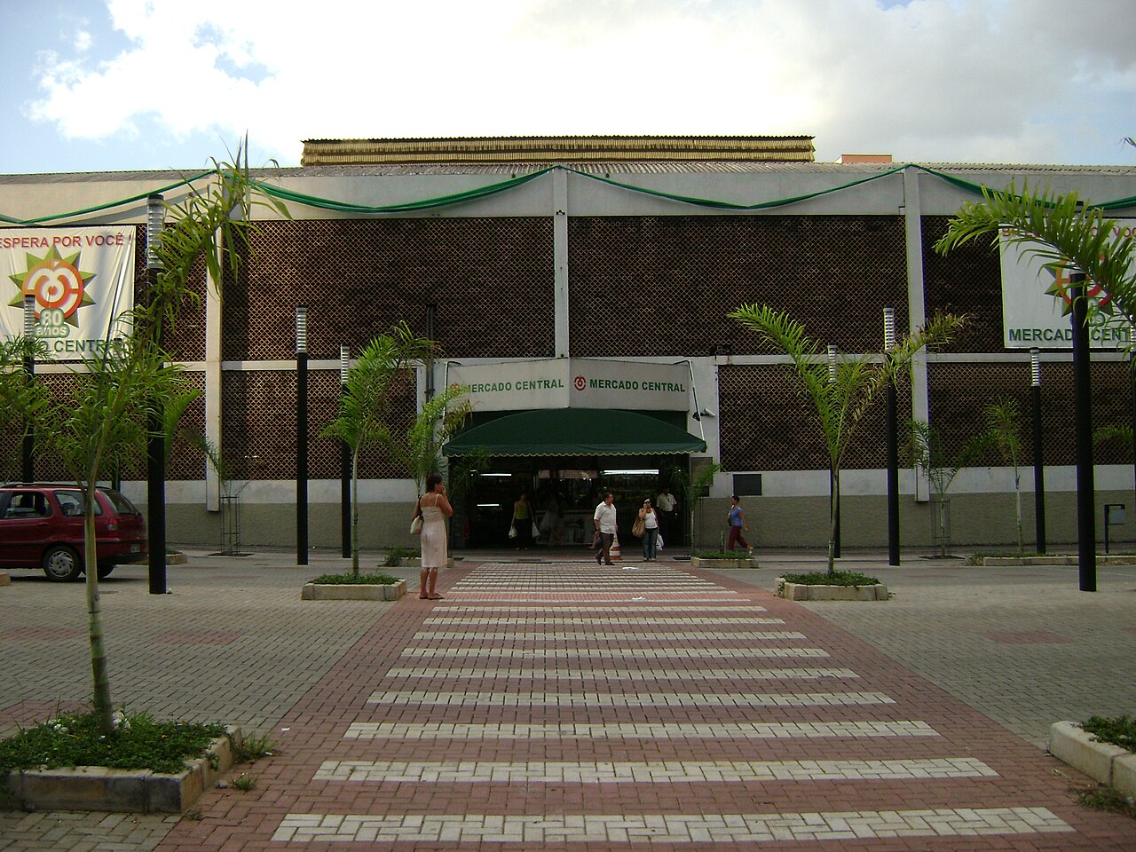 Fachada do Mercado Central com letreiros, entrada sob toldo verde, pessoas junto ao acesso e palmeiras.