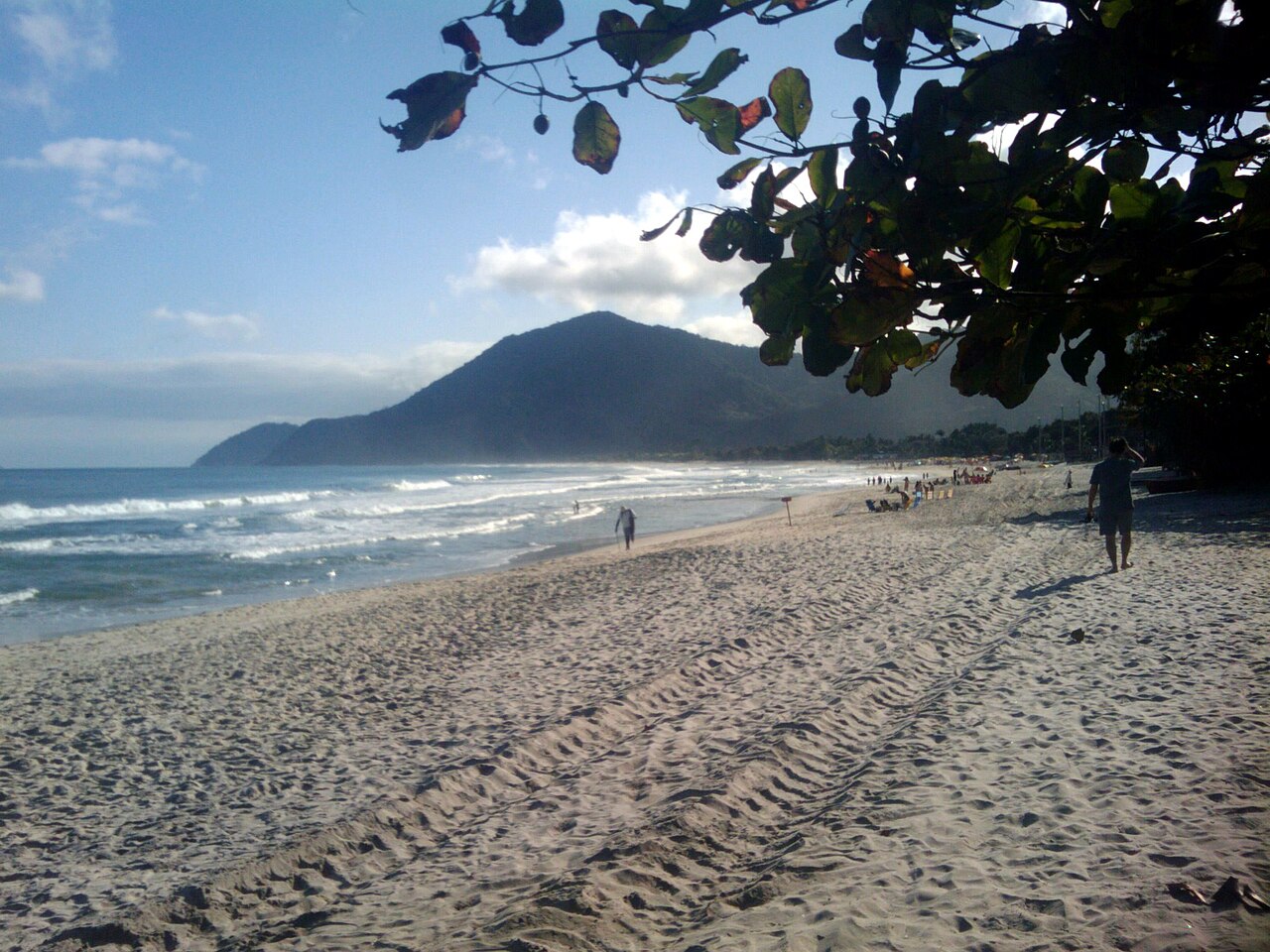 Praia de Maresias com areia larga marcada por pegadas e pneus, ondas, pessoas dispersas, morro vegetado e galhos no canto superior direito.