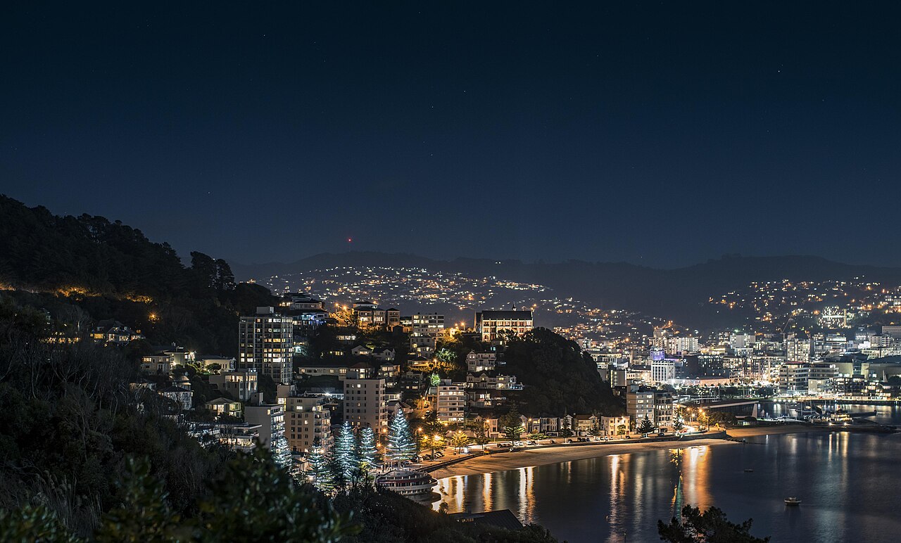 Vista noturna de Oriental Bay, com praia e avenida em curva, edifícios iluminados e reflexos na água.