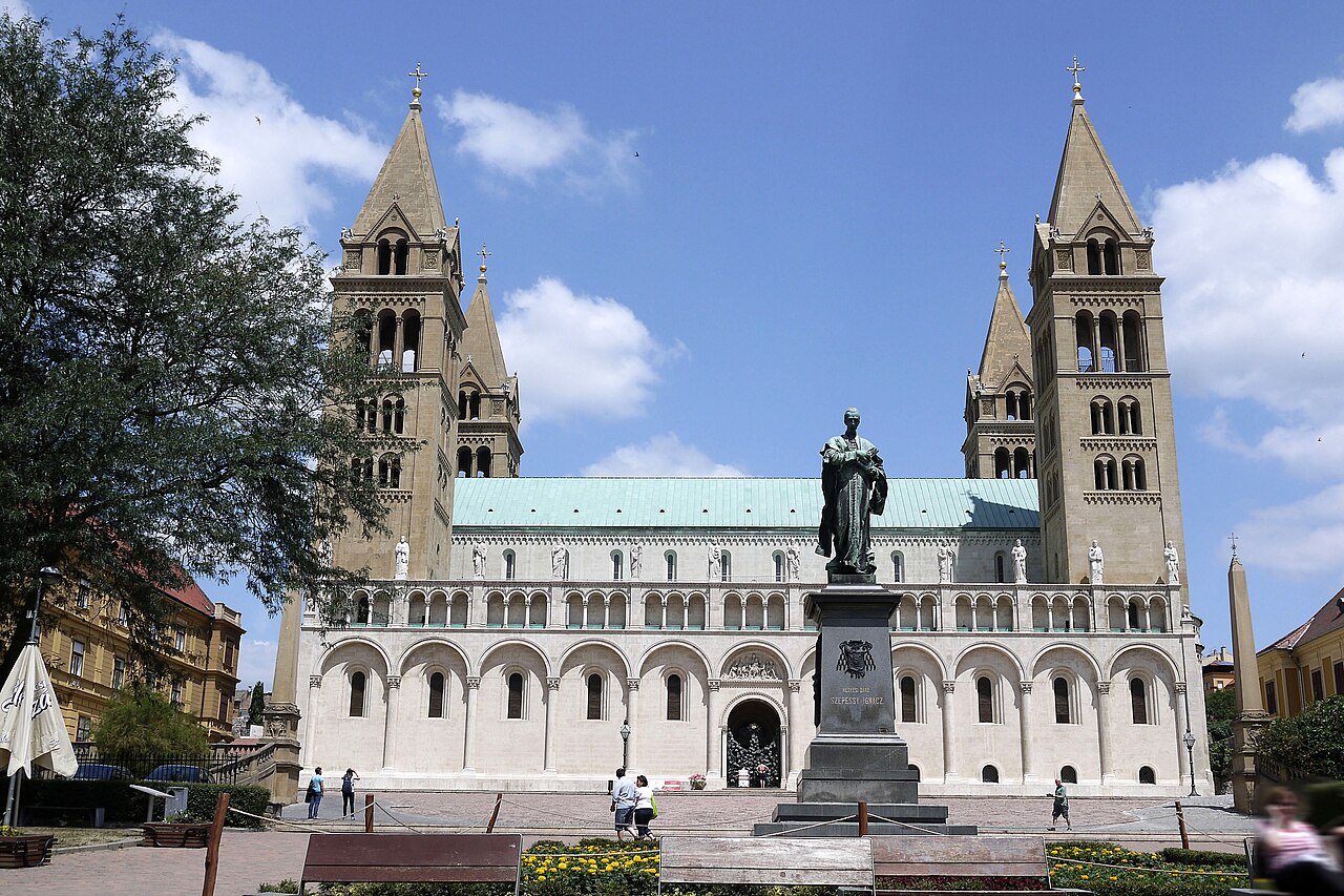 Exterior da Catedral de Pécs, com quatro torres pontiagudas, praça, bancos, árvores e pedestres.