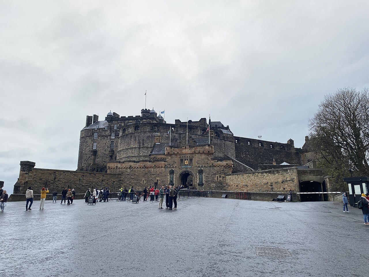 Muralhas e torres de pedra do Castelo de Edimburgo, com entrada em arco, visitantes e barreiras na esplanada sob céu encoberto.