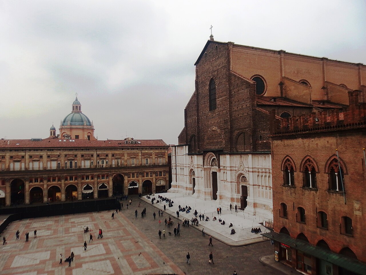 Vista elevada da Piazza Maggiore, com pavimentação geométrica, pedestres, edifícios com arcadas e a fachada de San Petronio à direita.