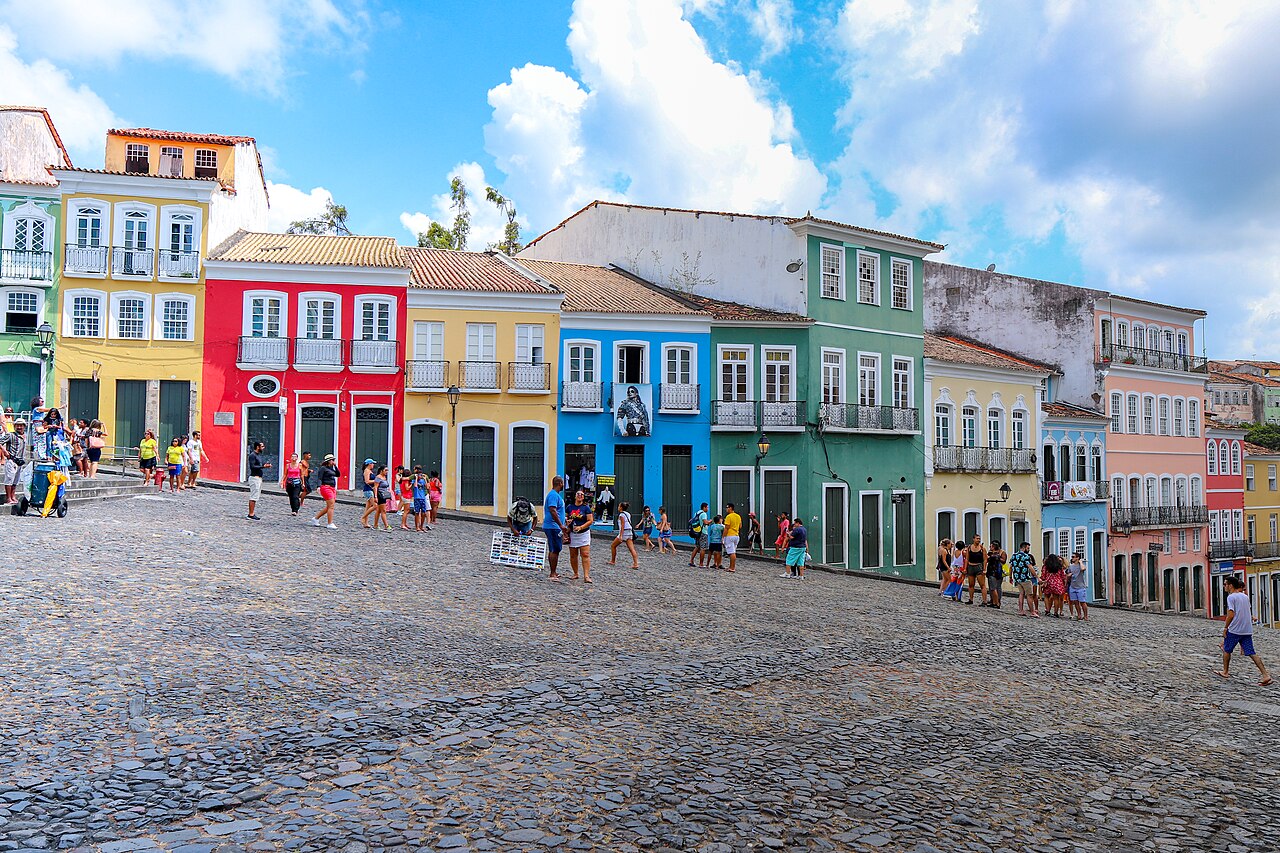 Largo do Pelourinho com piso de pedras em declive, pedestres e sobrados de fachadas coloridas sob céu azul com nuvens.
