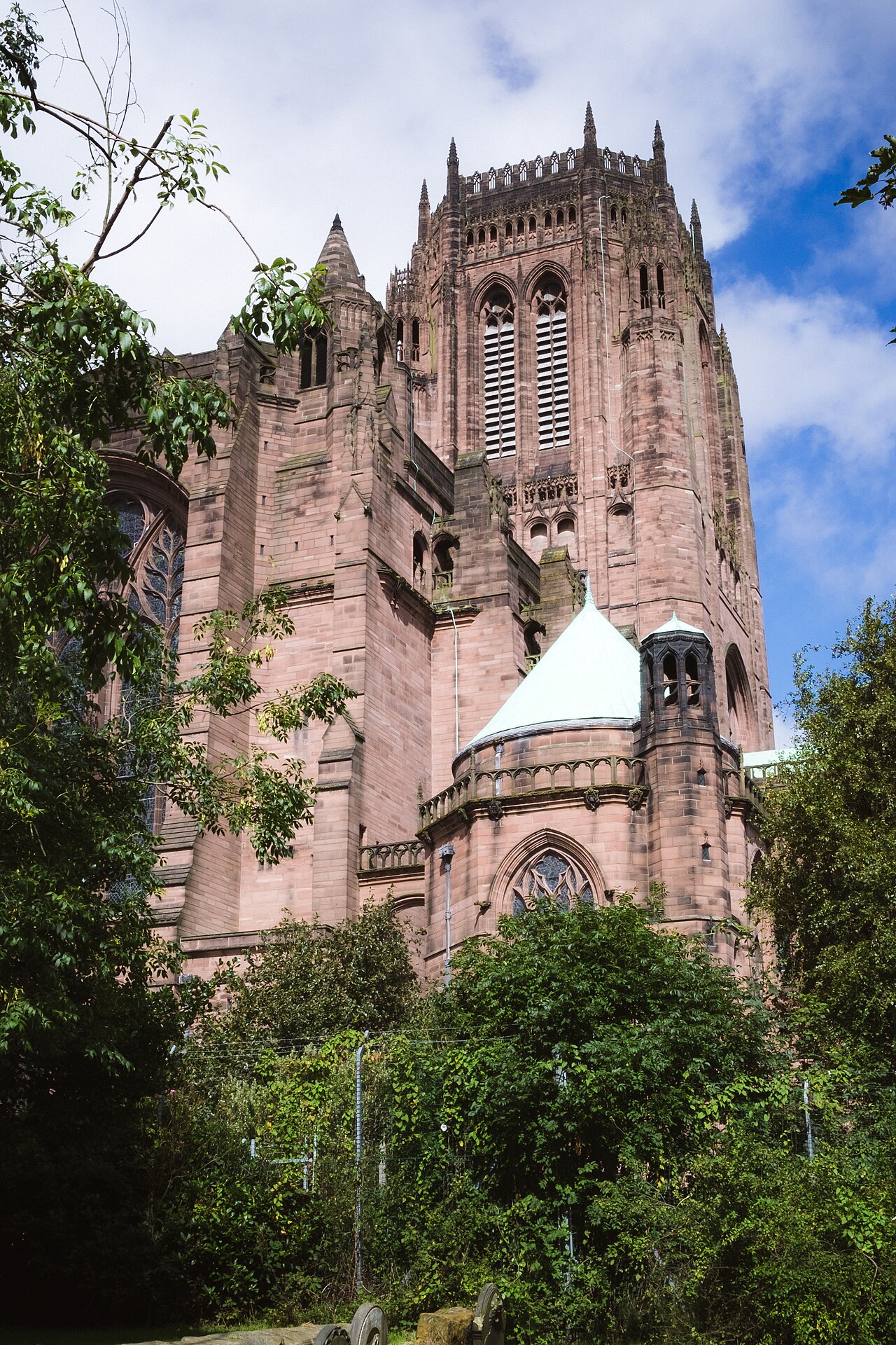 Exterior da Liverpool Cathedral visto de baixo, com torre quadrangular de pedra avermelhada, janelas ogivais, árvores, cerca e lápides em primeiro plano.