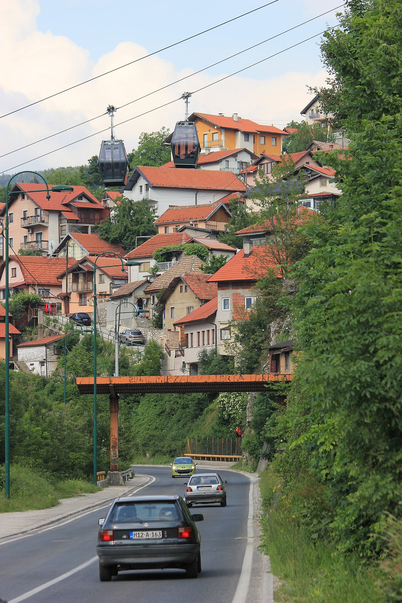 Duas cabines de teleférico sobre uma encosta com casas de telhados vermelhos, vegetação e uma estrada curva em Sarajevo.