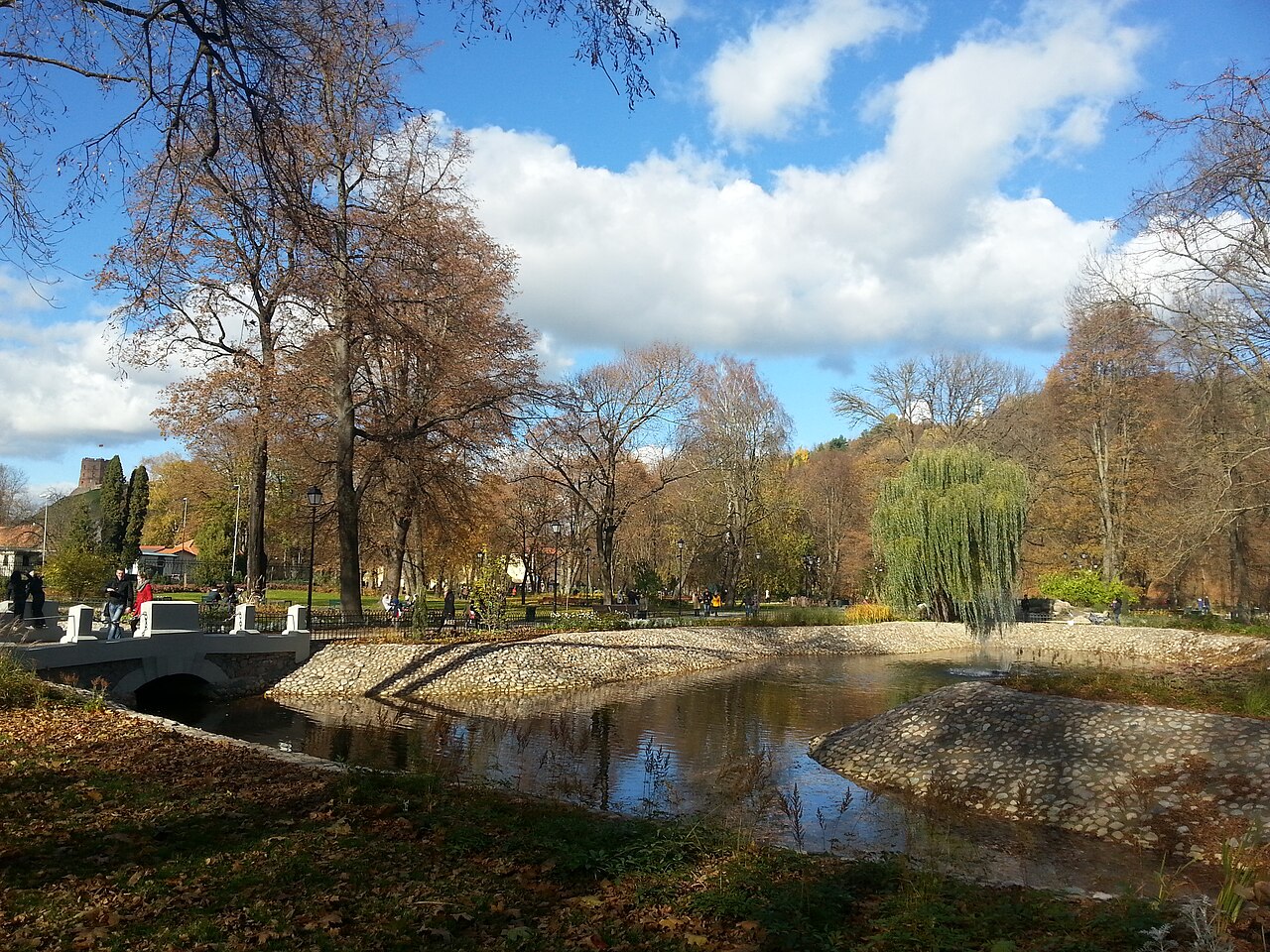 Jardim com árvores de folhagem outonal, lago de margens de pedra, pequeno jato de água, ponte baixa e visitantes nos caminhos.