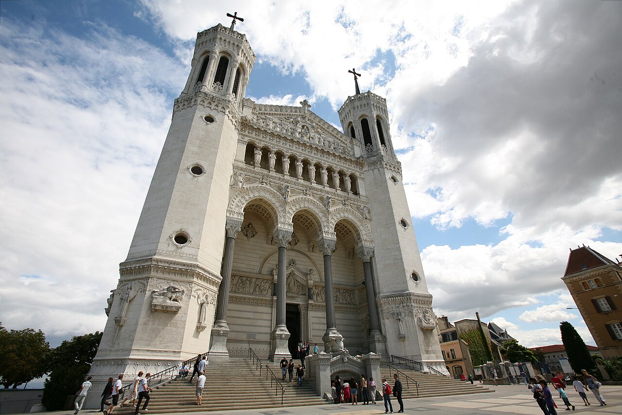 Fachada clara da basílica de Fourvière, vista de baixo, com duas torres, três grandes arcos e visitantes na escadaria.