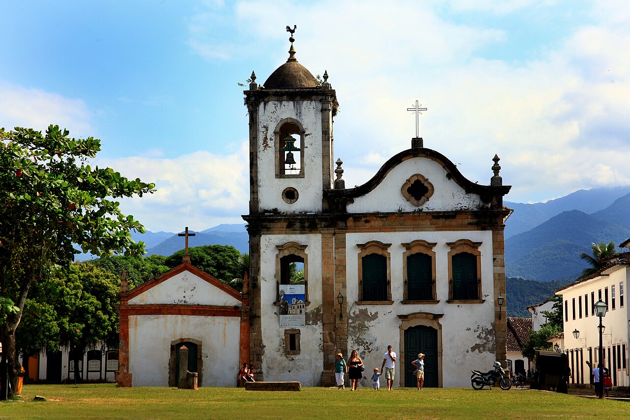 Igreja de Santa Rita branca, com detalhes de pedra, torre sineira à esquerda e portas escuras, diante de uma área gramada.